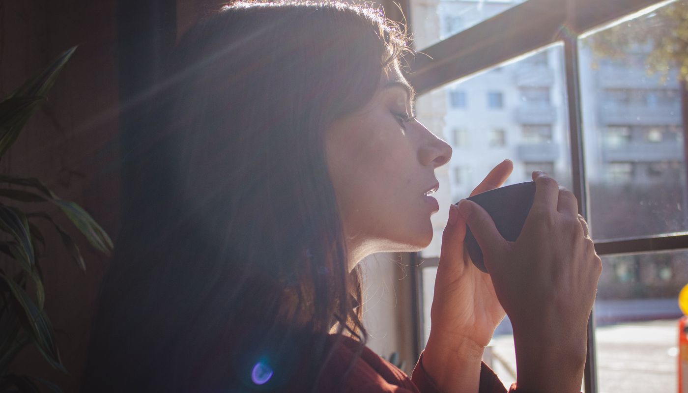 A woman pauses with a warm drink by a sunlit window